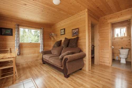 Cozy wooden cabin interior featuring a sofa, dining table, and a bathroom door in the background. Natural light from windows.