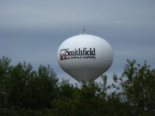 A white water tower with the text "Smithfield: ham, history & hospitality" surrounded by green trees under a cloudy sky.