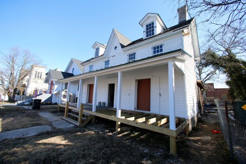 A white, two-story house with a porch and gabled roof, partially under renovation, set against a clear blue sky.