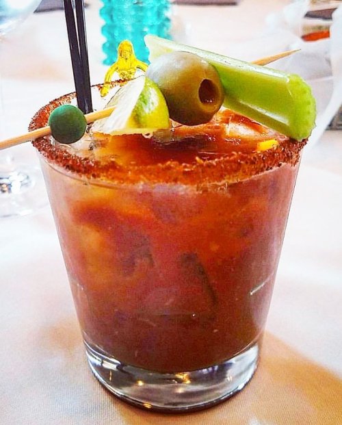 A close-up of a garnished cocktail with a salted rim, featuring olives, celery, and lime on a table setting.