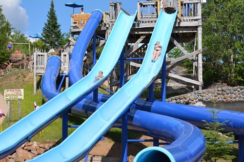 Two people slide down blue water slides at a park, with a wooden structure and greenery in the background.
