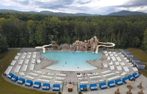 Aerial view of a large pool area with water slides, surrounded by lounge chairs and lush green trees in the background.
