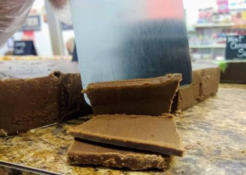 A hand using a metal spatula to cut a thick piece of fudge on a countertop, with some pieces already cut.