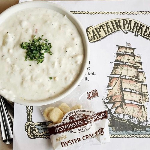 A bowl of clam chowder topped with herbs, accompanied by a pack of oyster crackers, on a nautical-themed placemat.