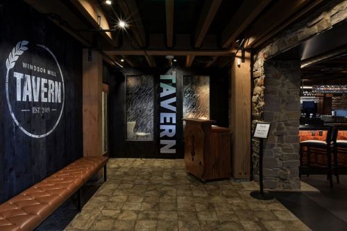 Interior of a tavern featuring a wooden reception area, stone walls, and a large "TAVERN" sign. Warm lighting enhances the ambiance.