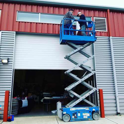 Two workers on a blue scissor lift are installing a window above a large garage door on a red metal building.