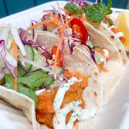 Three tacos filled with crispy fish, topped with fresh vegetables, herbs, and a drizzle of creamy sauce, served with lemon.