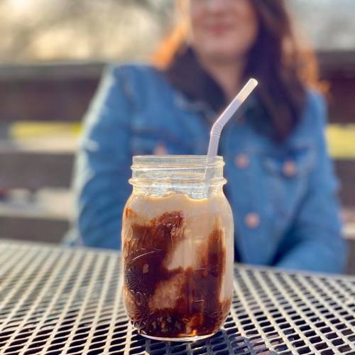 A jar of iced coffee with cream and a straw, set on a table with a blurred figure in the background.