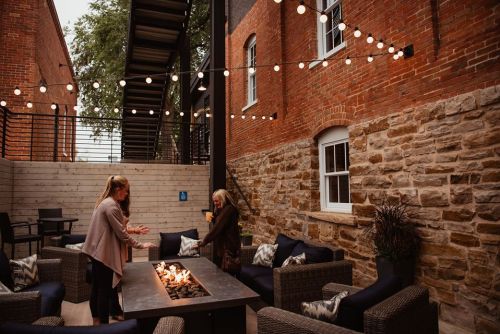 Two women converse near a fire pit in a cozy outdoor space, surrounded by string lights and brick walls.