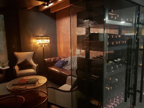 Cozy lounge area with a leather sofa, elegant lighting, and a glass wine cabinet filled with bottles.