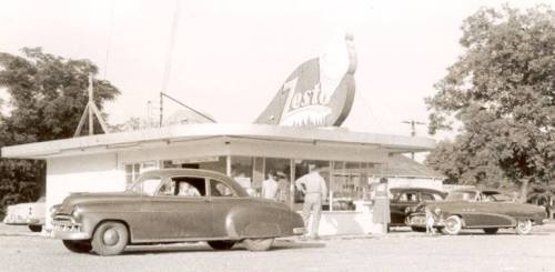 A vintage drive-in diner with classic cars parked outside, featuring a large Zesto sign and people at the counter.