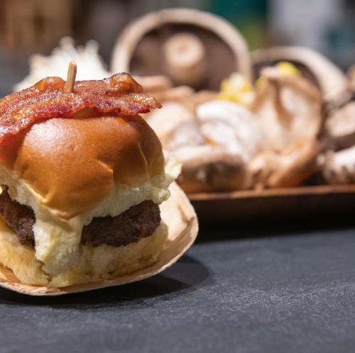 A close-up of a mini burger topped with bacon, set against a blurred background of assorted mushrooms.