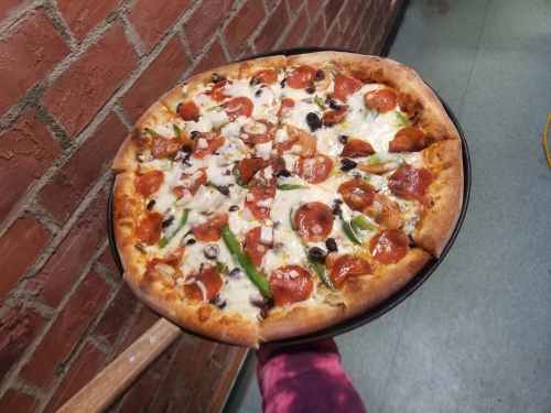 A person holds a large pizza topped with pepperoni, olives, green peppers, onions, and cheese on a black tray.