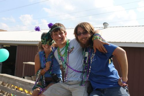 Three friends celebrate together, wearing colorful beads and masks, smiling and enjoying a festive outdoor event.
