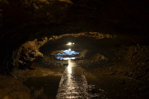A dimly lit cave tunnel with a wet path and rocky walls, leading to a distant light at the end.