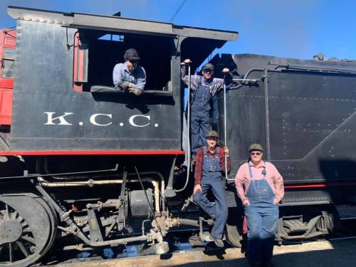 Four people in vintage work attire pose by a black steam train, with one inside the cab and three outside.