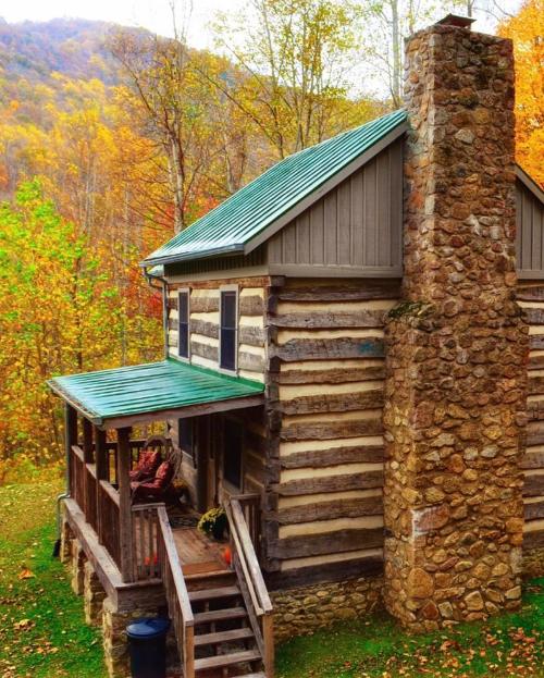 A rustic log cabin with a stone chimney, surrounded by vibrant autumn foliage and a green metal roof.