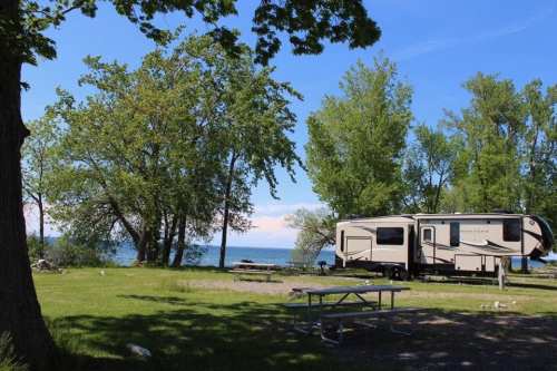 A camper parked by a lake, surrounded by green trees and a clear blue sky. A picnic table is nearby.