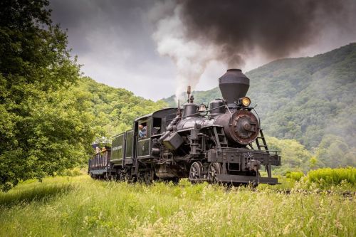 A vintage steam locomotive chugs through a lush green landscape, billowing smoke against a backdrop of rolling hills.