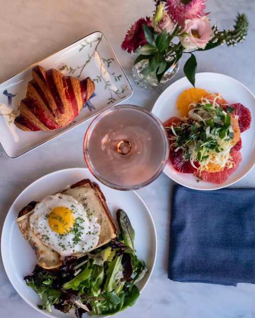 A table setting with a croissant, a salad with an egg, and a citrus salad, accompanied by a glass of rosé and flowers.