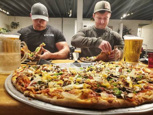 Two men sit at a table with a large pizza and beers, focused on their meals in a casual dining setting.