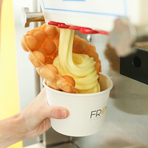 A hand holds a cup as soft serve ice cream is dispensed into it, accompanied by a bubble waffle.