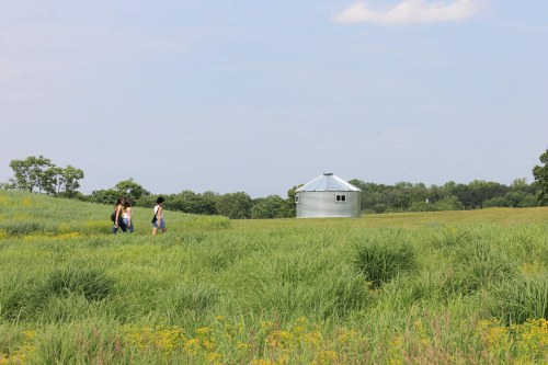 Two women walk through a grassy field towards a silver grain silo under a clear blue sky.