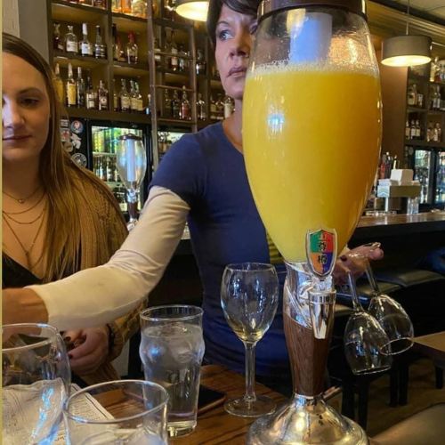 A woman pours a large glass of orange drink at a bar, with another woman seated nearby and various glasses on the table.
