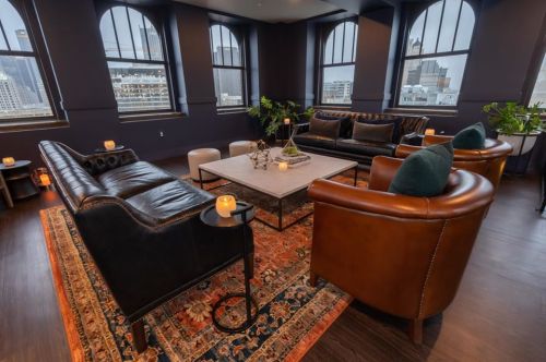 Stylish lounge with leather sofas, a coffee table, and city views through large windows, accented by plants and candles.