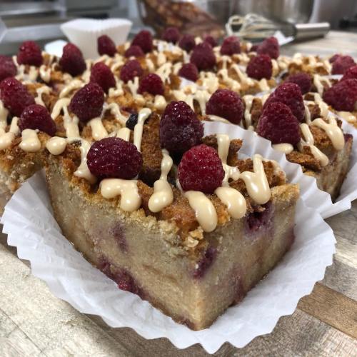 A close-up of a square dessert topped with raspberries and drizzled with a creamy sauce, served in a paper liner.