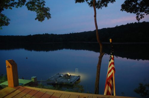 A serene lake at dusk, with a dock illuminated by a light and an American flag in the foreground.