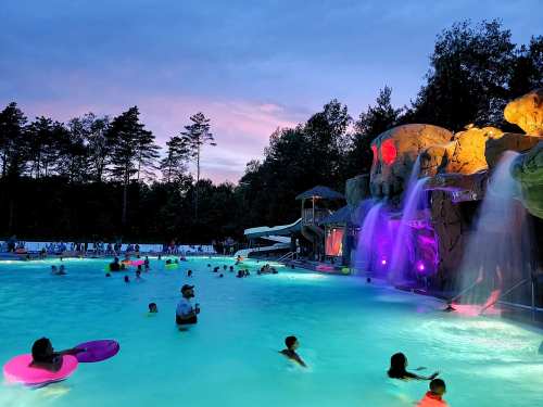 A vibrant pool scene at dusk, featuring a waterfall, slides, and people enjoying the water under colorful lights.