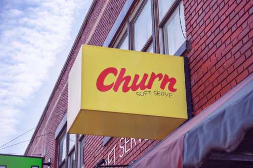 Sign for "Churn Soft Serve" in bold red letters on a bright yellow background, mounted on a brick building.