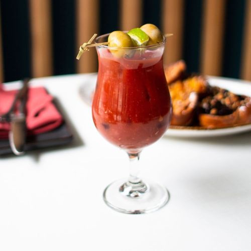A vibrant red cocktail garnished with olives and lime, served in a tall glass on a table with food in the background.