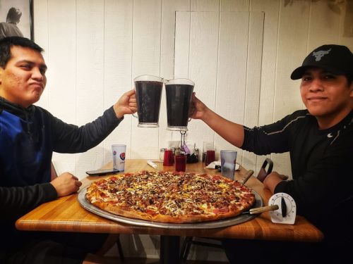 Two men clink large glasses of dark drinks over a large pizza on a table, smiling at each other.