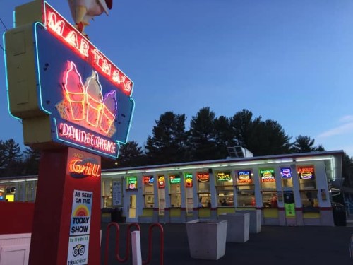 Neon sign for Martha's Dairy Bar and Grill at dusk, with food stalls and trees in the background.