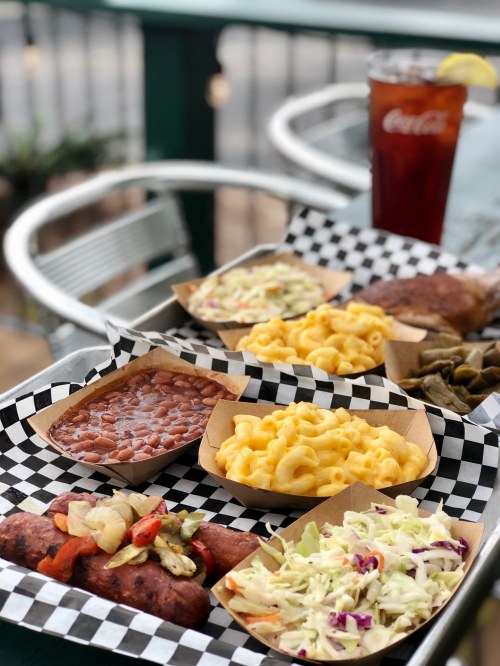 A tray of food featuring a sausage, baked beans, macaroni and cheese, coleslaw, and pickled vegetables, with a drink.
