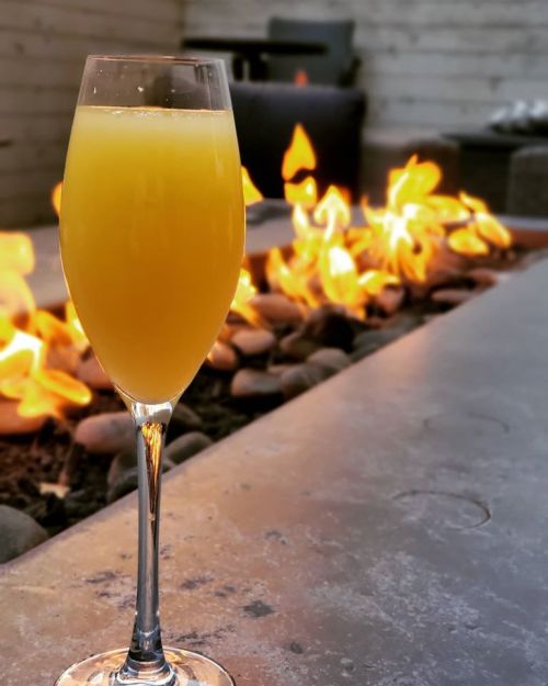 A glass of orange mimosa stands beside a glowing fire pit with smooth stones.