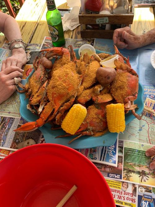 A blue platter filled with seasoned crabs, potatoes, and corn, surrounded by hands and a green bottle on a table.