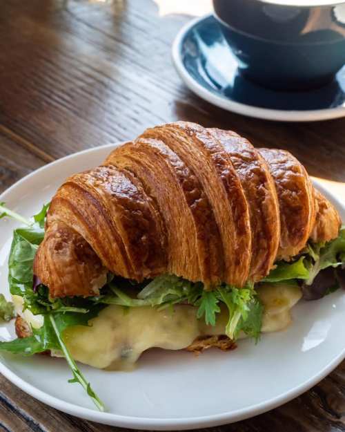 A golden croissant sandwich filled with greens and melted cheese, served on a white plate with a coffee in the background.