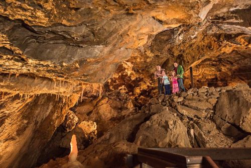 A group of people stands on a rocky ledge inside a cave, surrounded by stalactites and illuminated by warm lighting.