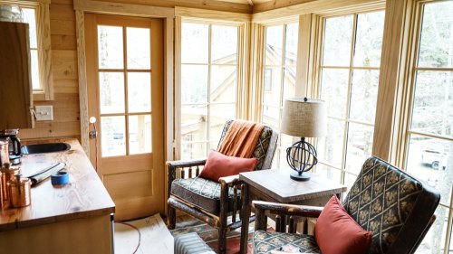 Cozy sunlit room with two chairs, a table, and large windows overlooking a natural setting.