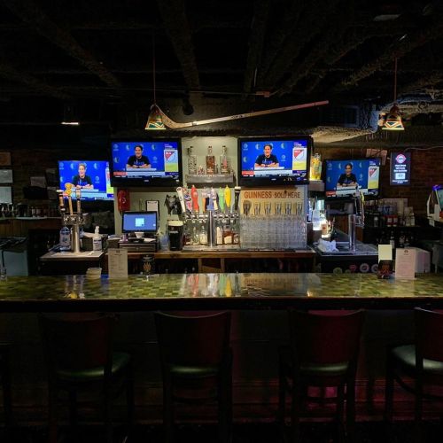 A dimly lit bar with a long counter, multiple TVs showing sports, and a variety of drinks displayed behind the bar.