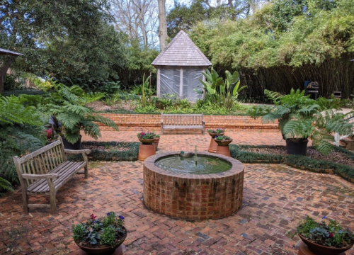 A serene garden with a brick patio, a circular fountain, benches, and lush greenery surrounding a gazebo.