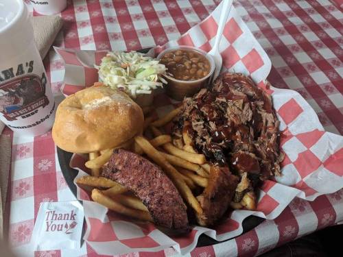 A plate of barbecue with pulled meat, fries, coleslaw, baked beans, a roll, and a drink on a checkered tablecloth.