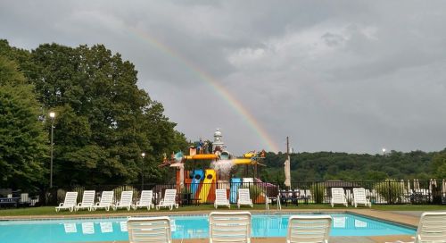 A colorful water play area with a rainbow in the background, viewed from a poolside with lounge chairs.