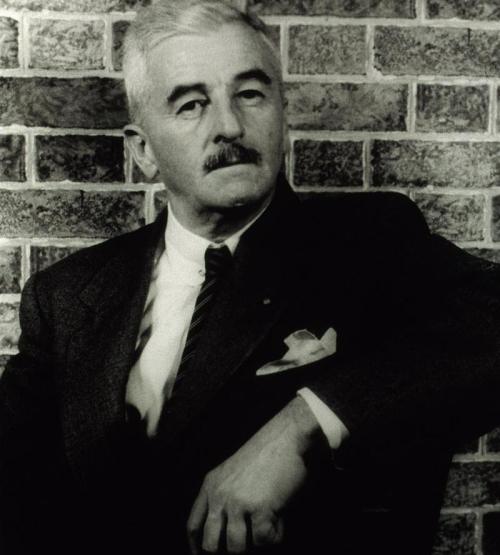 A black-and-white portrait of a distinguished man with a mustache, dressed in a suit, seated against a brick wall.