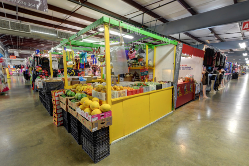 A colorful market stall filled with fresh fruits, surrounded by various shops in a spacious indoor market.