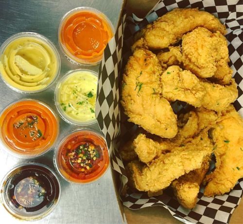 A box of crispy fried chicken tenders with six assorted dipping sauces in plastic cups.