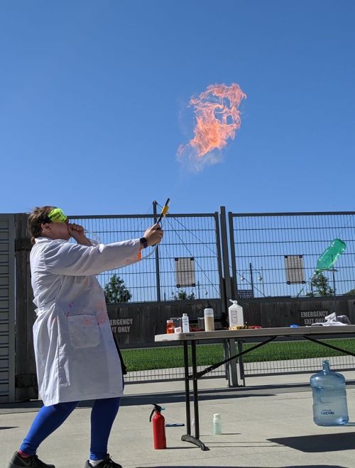 A person in a lab coat and goggles ignites a flame outdoors, with a clear blue sky in the background.